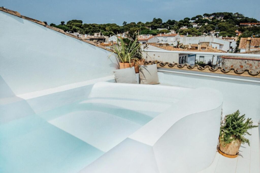 A small white rooftop pool with clear water is surrounded by potted plants and cushions, offering a scene that rivals boutique hotels in Costa Brava—overlooking a town of white buildings and green trees beneath a blue sky.