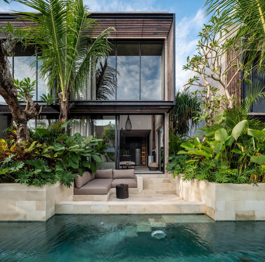 14 UNIQUE Boutique Hotels in Canggu, Bali 50 Modern villa with large glass windows, surrounded by lush tropical plants, featuring an outdoor seating area and steps leading to a private swimming pool—an oasis reminiscent of boutique hotels in Canggu under a blue sky.