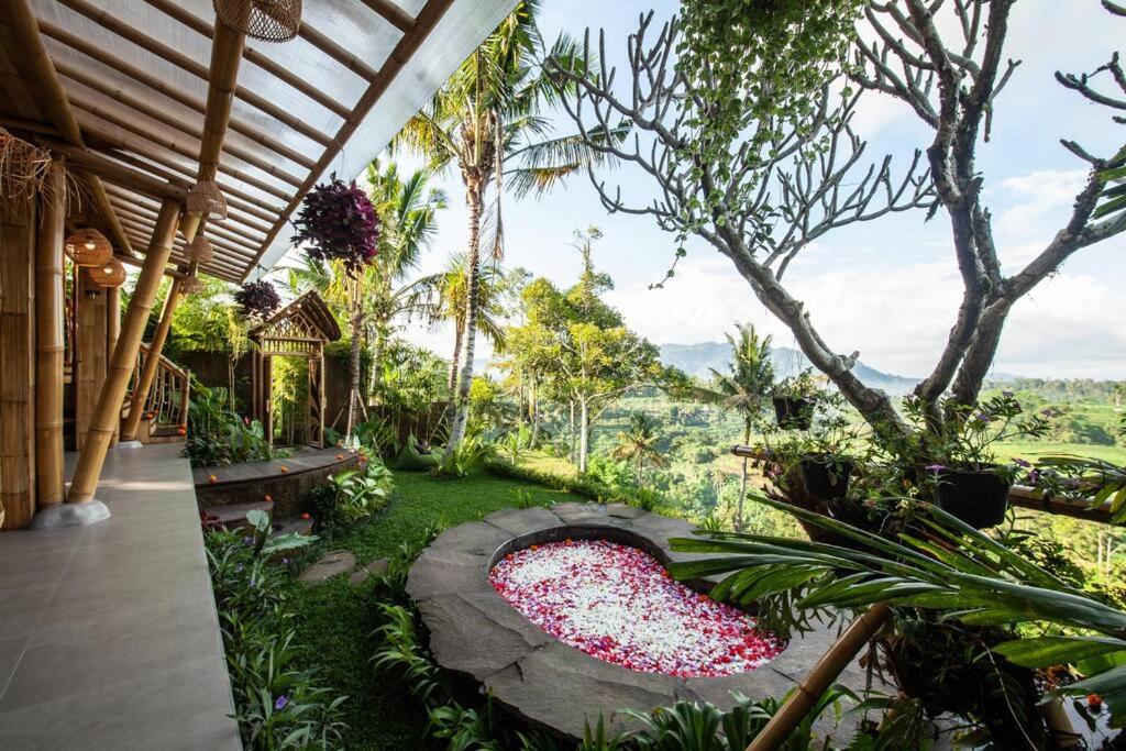 A tropical outdoor patio at one of the best family villas in Bali, featuring a circular stone hot tub filled with flower petals, lush greenery, bamboo structures, and stunning views of distant mountains and rice fields.