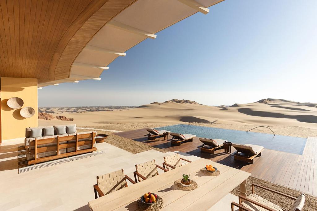 9 Best Hotels in Saudi Arabia: Unique Stays Across the Country 26 A luxurious outdoor patio at one of the Best Hotels in Saudi Arabia features modern wooden furniture, a dining table, lounge chairs, and an infinity pool overlooking vast sand dunes under a clear blue sky.