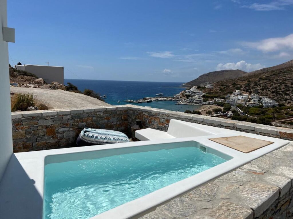 Authentic Hotels in Sikinos, Cyclades 1 A small private pool with clear blue water overlooks a coastal village with white buildings, hills, and the blue sea under a partly cloudy sky—an ideal escape for guests seeking hotels in Sikinos. Stone walls and a lounge area surround the pool.
