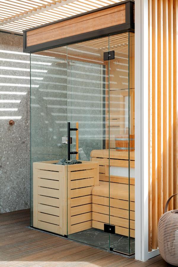 Where to Stay in Madrid: 14 Best Hotels 59 A modern glass-enclosed sauna with wooden benches and walls, featuring a heater in the corner, is illuminated by natural light streaming through slatted wooden panels—an amenity to consider when searching for where to stay in Madrid first time.