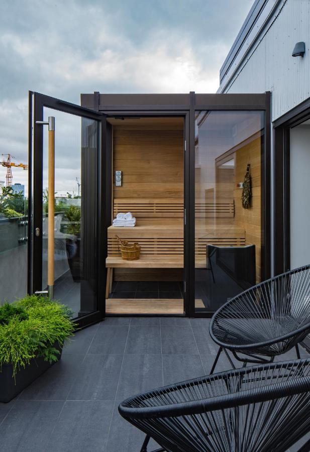 Where to Stay in Reykjavík: Top Guide 16 A modern outdoor sauna with glass doors and a wooden interior sits on a rooftop patio—an ideal spot for those searching where to stay in Reykjavik. Black chairs and green plants decorate the tiled area, set against a backdrop of cloudy sky.