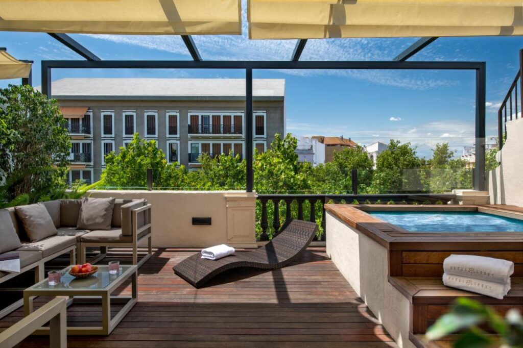 10 UNIQUE Hotels with Pool in Seville 4 A rooftop terrace with outdoor seating, a lounge chair, and a hot tub overlooks green trees and a building with balconies under a sunny, blue sky—perfect for those seeking hotels with pool in Seville.