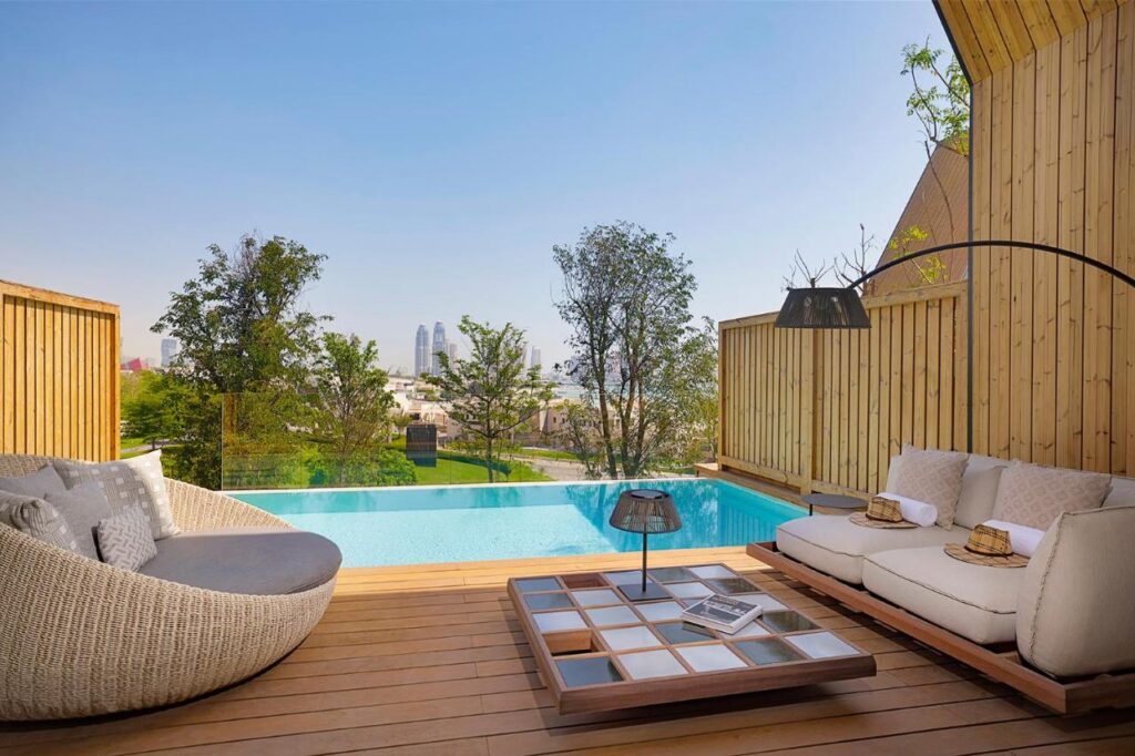 Where to Stay in Doha, Qatar: Transit, City & Beach Hotels 9 A stylish outdoor patio with a sofa, lounge chair, and low table overlooks a private infinity pool—an inviting option for those seeking where to stay in Doha. Wooden fencing and trees offer privacy, while the cityscape shimmers under a clear sky.