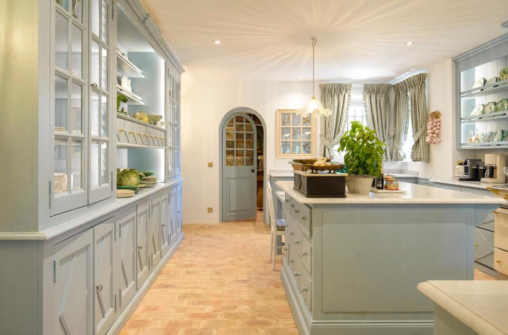 11 Unique Places to Stay in Provence 28 A spacious, light-filled kitchen with light blue cabinets, a central island, open shelving, checkered curtains, a potted plant on the counter, and a brick-patterned floor—evoking the charm of unique places to stay in Provence. Cozy and inviting throughout.