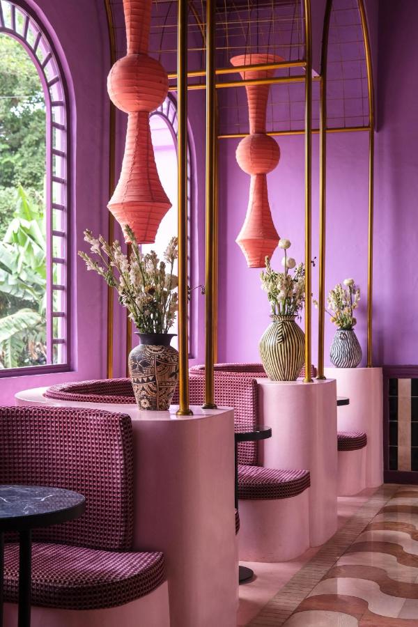 14 UNIQUE Boutique Hotels in Canggu, Bali 47 A vibrant cafe interior reminiscent of boutique hotels in Canggu, with pink and purple walls, patterned curved seating booths, black tables, large windows, and decorative vases filled with flowers. Pink paper lanterns hang from the gold-framed ceiling.