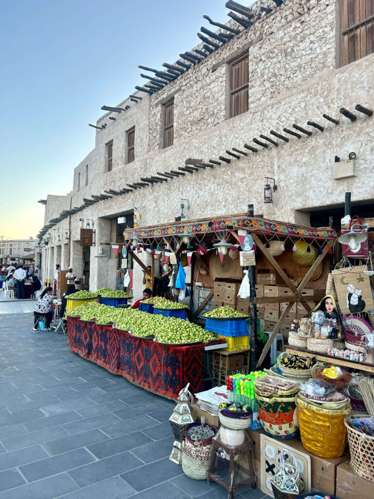 A bustling outdoor market with colorful fruit stands, baskets, and traditional woven items sits in front of a rustic stone building—perfect for travelers seeking authentic experiences while exploring where to stay in Doha. People are visible in the background under a clear sky.