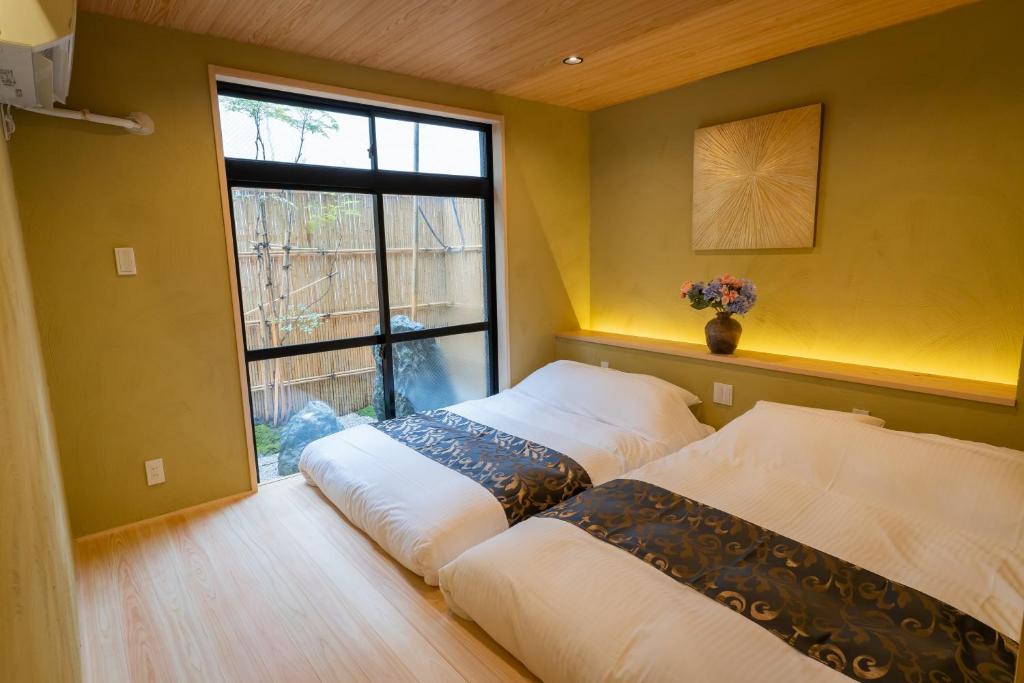 8 BEST Ryokans in Osaka 3 A cozy bedroom reminiscent of ryokans in Osaka, with two low, side-by-side beds covered in white linens and decorative runners, a vase of flowers on a shelf, a painting above, and a large window overlooking a small garden.