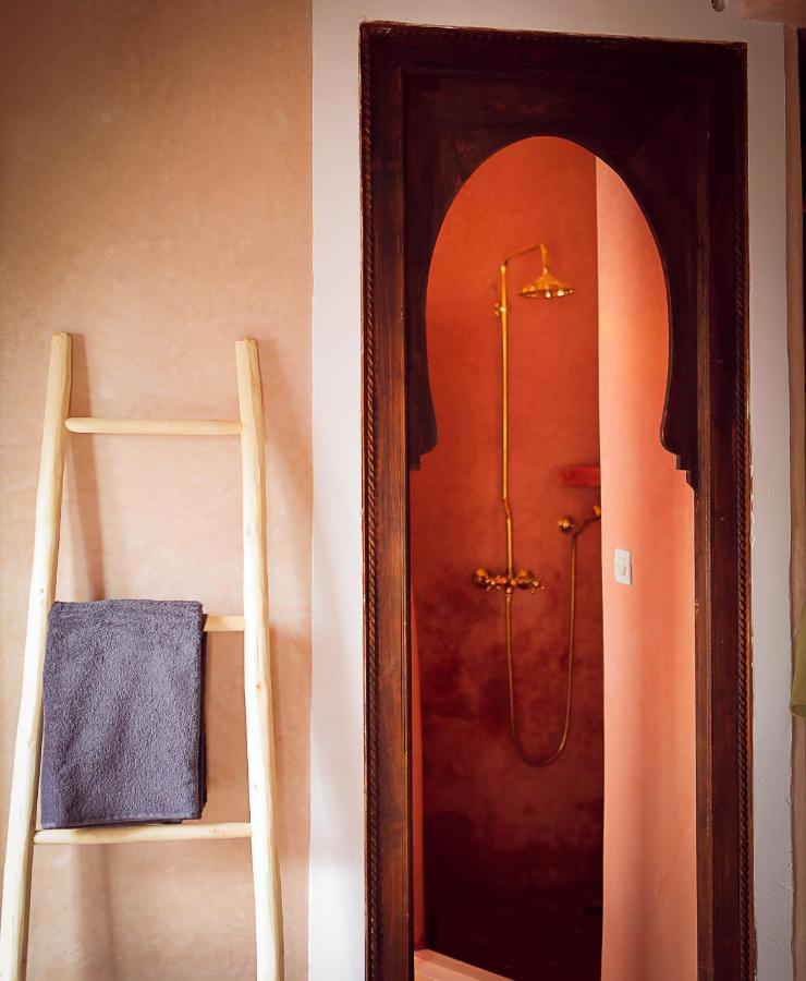 12 Charming Budget Riads in Marrakech 17 A wooden ladder with a folded gray towel leans against a pink wall next to a dark wooden arched doorway, revealing a shower with gold fixtures inside a softly lit bathroom, evoking the charm of budget riads in Marrakech.