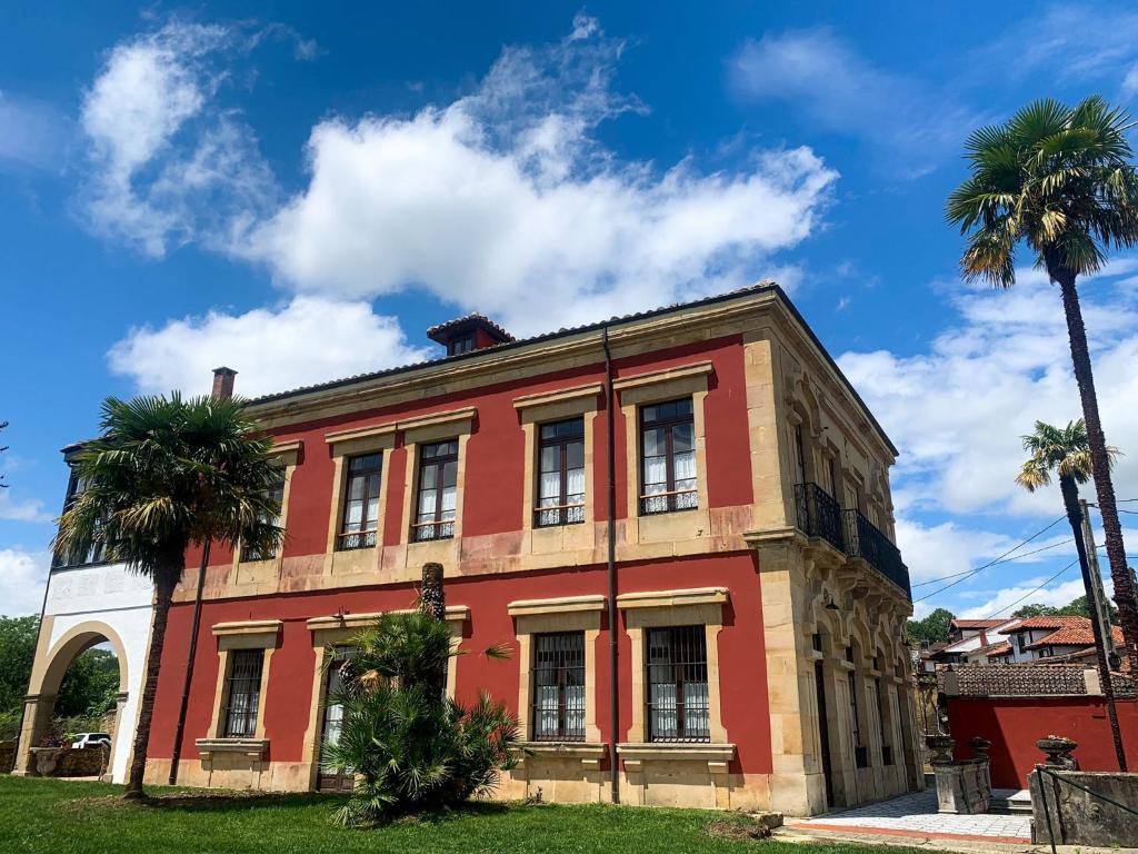 A large two-story red and beige building with tall windows, surrounded by palm trees and a blue sky with scattered clouds—reminiscent of the charming boutique hotels in Asturias.