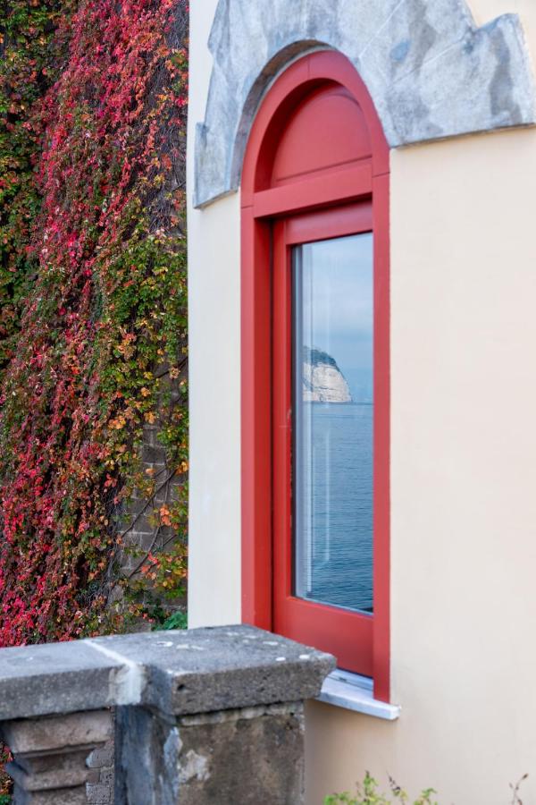 Boutique Hotels on the Amalfi Coast 8 A window with a red frame in a cream-colored wall reflects a distant rocky cliff by the sea, evoking the charm of boutique hotels on the Amalfi Coast. Red and green ivy climbs the adjacent wall, adding color to the scene.