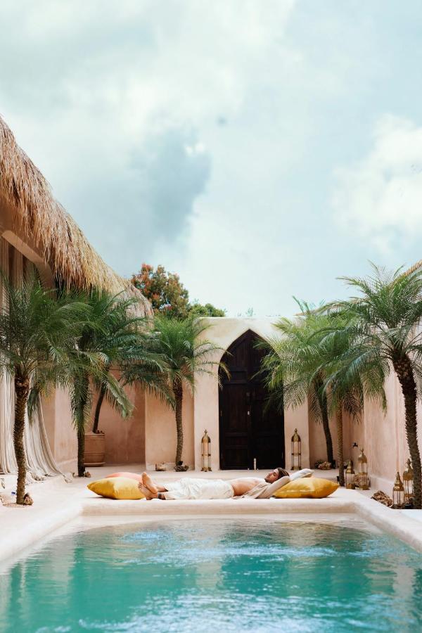 A serene poolside scene at one of the best family villas in Bali, featuring two people relaxing on large yellow cushions beside a turquoise pool, surrounded by palm trees and beige stucco walls under a partly cloudy sky.