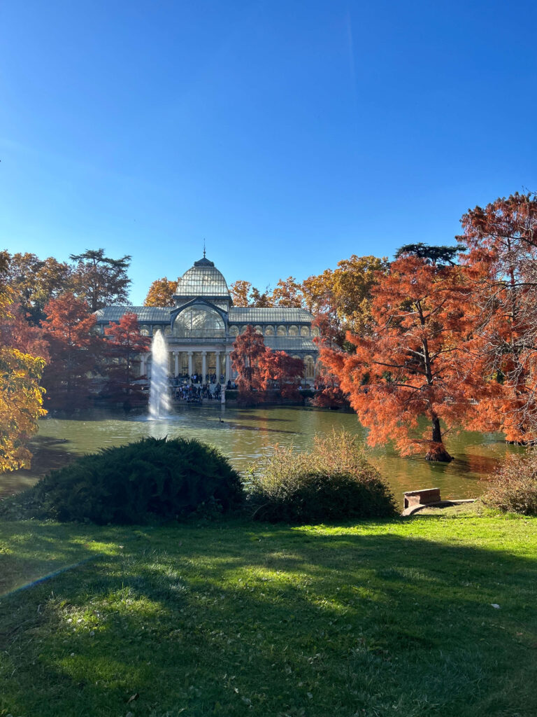 Where to Stay in Madrid: 14 Best Hotels 60 Glass-domed building by a pond with a fountain, surrounded by trees in autumn hues of red, orange, and yellow under a clear blue sky—an idyllic spot for those exploring where to stay in Madrid first time. Sunlight brightens the green grass nearby.