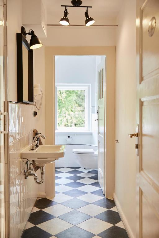 FAIRYTALE Castle Hotels in Bavaria 5 A narrow bathroom with black and white checkered tile floor, wall-mounted sink, mirror, and lights—evoking the elegance of castle hotels in Bavaria—leads to a toilet by a window with greenery visible outside.