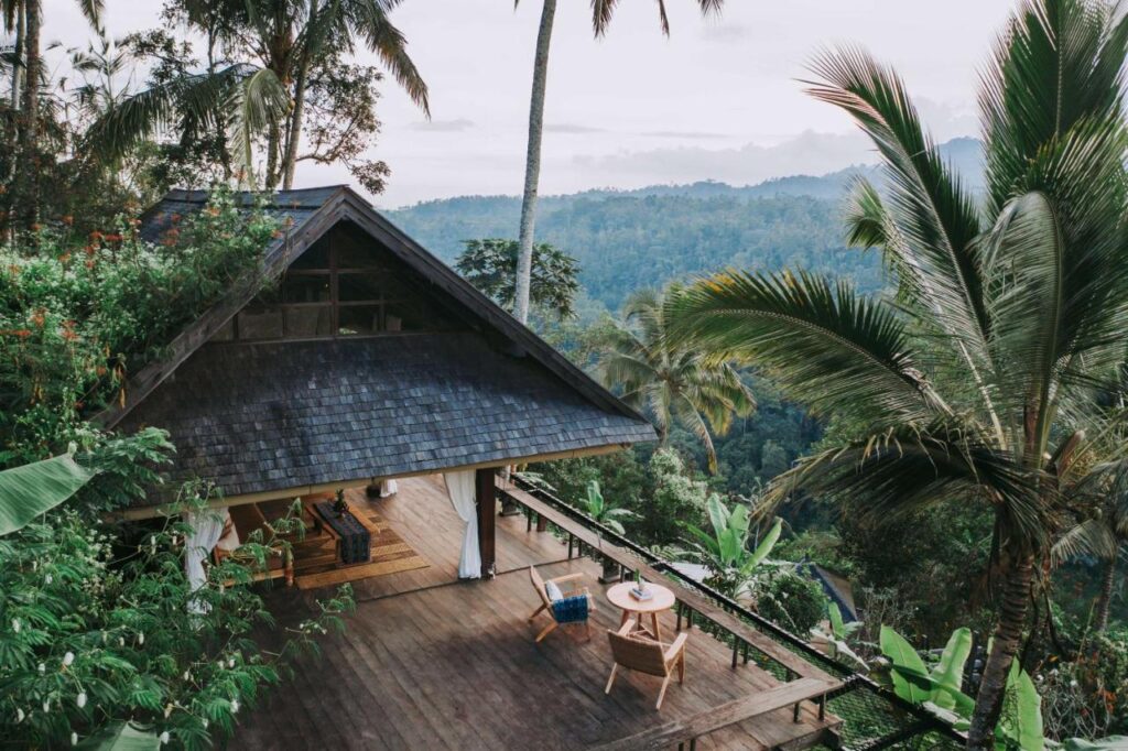 13 Most UNIQUE Places to Stay in Ubud 16 A wooden villa with a thatched roof sits amidst lush tropical trees, featuring an open-air patio with chairs and a table—one of the unique places to stay in Ubud—overlooking a green jungle and distant hills under a cloudy sky.