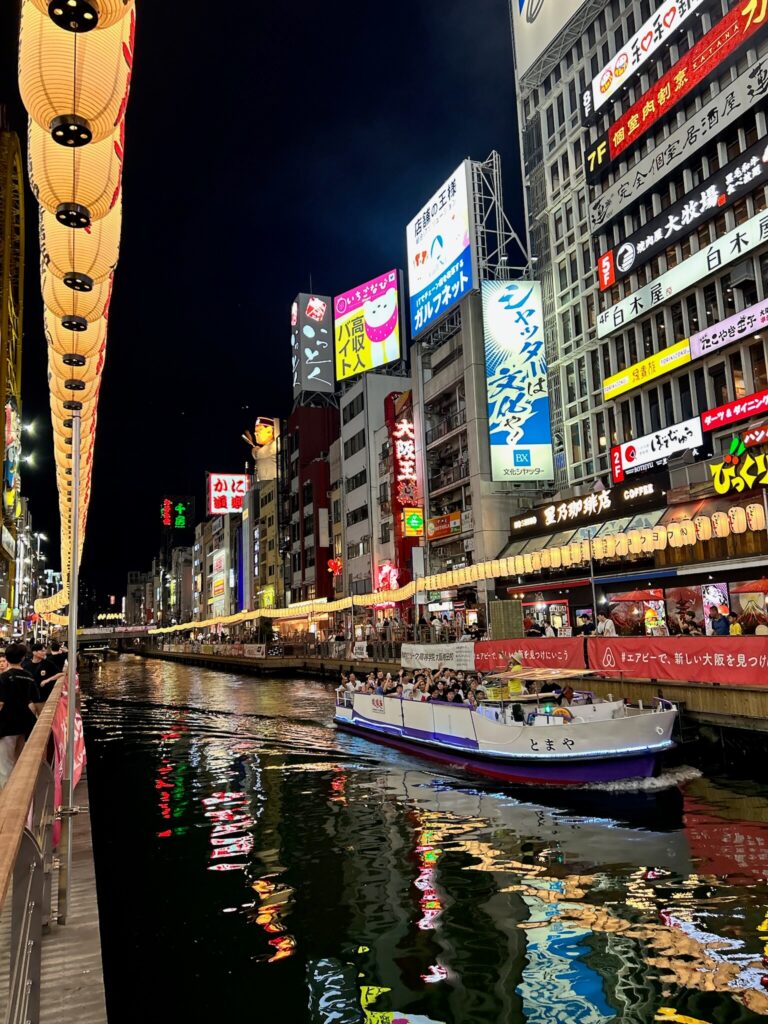8 BEST Ryokans in Osaka 38 A brightly lit canal at night in a bustling city with colorful neon signs, lanterns, and advertisements. A boat cruises along the water by vibrant crowds and riverside ryokans in Osaka, all reflected in the shimmering city lights.
