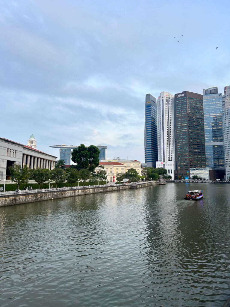 13 Most Unique Hotels in Singapore for an Incredible Holiday 56 A calm river flows through a city with modern skyscrapers, unique hotels in Singapore, and older classical buildings along the bank; a small boat with passengers floats on the water under a partly cloudy sky.