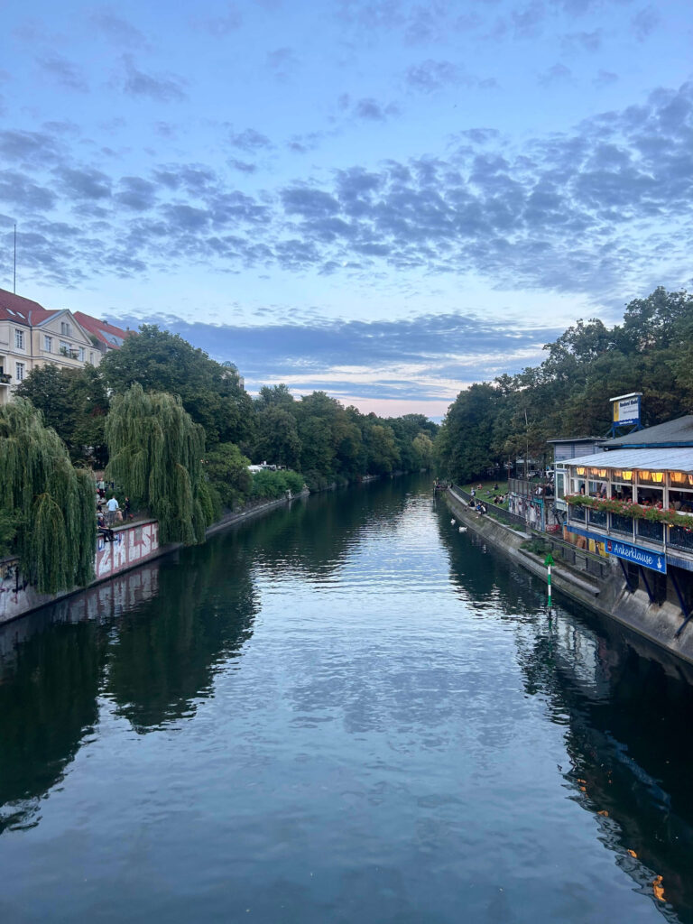 Boutique Hotels in Berlin Kreuzberg 3 A calm river lined with trees and buildings on both sides at dusk, with a partly cloudy sky above and reflections shimmering in the water. People gather along the riverside walkways near boutique hotels in Berlin Kreuzberg.