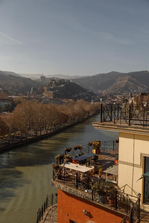 11 Unique Hotels in Georgia You Can't Miss! 23 A riverside view of a city with a terrace cafƩ in the foreground, lined with potted plants. Bare trees border the river, and hills dotted with unique hotels in Georgia rise in the background under a clear sky.