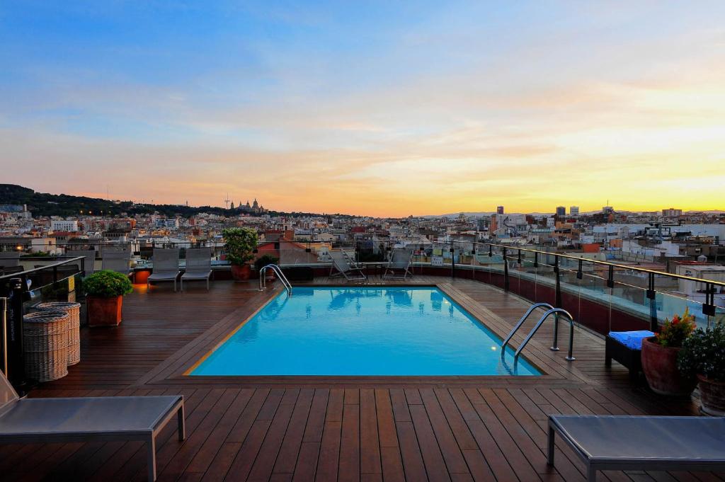 Top Barcelona Hotels with Rooftop Pool 34 Rooftop pool with wooden deck, lounge chairs, and potted plants overlooking a cityscape at sunset—perfect for guests seeking Barcelona Hotels with Rooftop Pool and breathtaking views of the colorful sky and distant buildings.