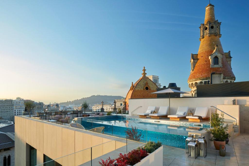 Top Barcelona Hotels with Rooftop Pool 29 Rooftop infinity pool with lounge chairs and umbrellas overlooking a cityscape with historic, turreted buildings under a clear blue sky at sunset—a signature feature of the best Barcelona hotels with rooftop pool.