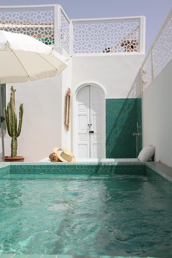 12 Charming Budget Riads in Marrakech 20 A serene outdoor pool with turquoise water is surrounded by white walls, a green-tiled accent wall, a tall cactus, rolled towels, a hat, and an umbrella—capturing the tranquil charm of budget riads in Marrakech. A white arched door is centered in the background.