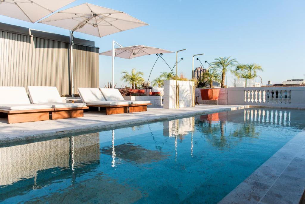 Top Barcelona Hotels with Rooftop Pool 49 A rooftop pool with clear blue water awaits at one of the top Barcelona hotels with rooftop pool, surrounded by white lounge chairs, large umbrellas, and potted palms under a clear blue sky.