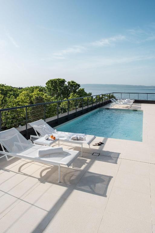 8 TOP Boutique Hotels in Split 27 A modern outdoor pool with clear blue water, surrounded by white lounge chairs and towels, overlooks lush green trees and a distant sea—an ideal spot for those seeking where to stay in Split under a clear sky.