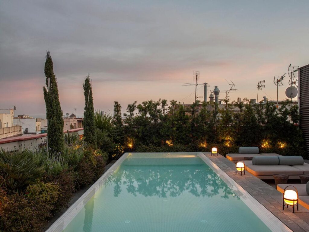 13 Unique Hotels in Barcelona, Spain 54 A rooftop pool surrounded by greenery and lounge chairs, softly lit by small lamps at dusk, with a city skyline and antennas visible—a relaxing scene typical of Barcelona Hotels with Rooftop Pool under a partly cloudy sky.