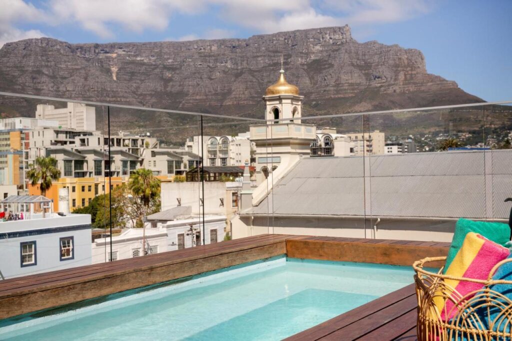 Unique Places to Stay in Cape Town 5 A rooftop pool with colorful towels on a lounge chair overlooks city buildings and a gold-domed tower, with a large mountain in the background—one of the unique places to stay in Cape Town under a partly cloudy sky.