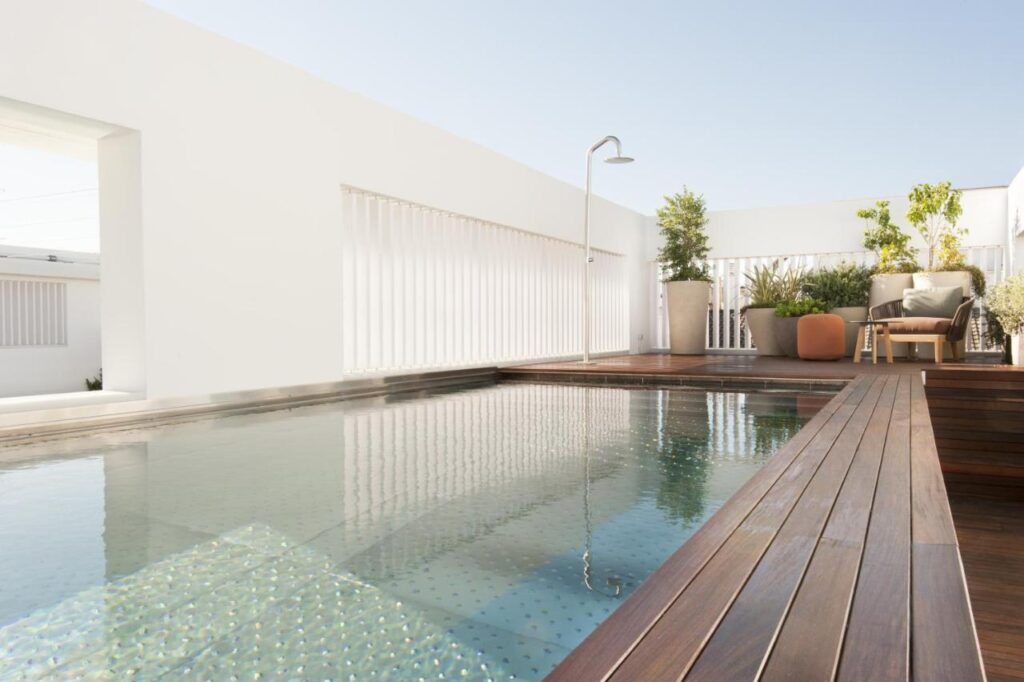 10 UNIQUE Hotels with Pool in Seville 17 A modern outdoor terrace at one of the hotels with pool in Seville, featuring a small rectangular pool, wooden decking, an outdoor shower, lush potted plants, and a cozy seating area beneath clear skies.