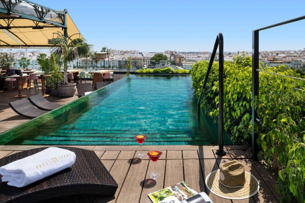 10 UNIQUE Hotels with Pool in Seville 2 A rooftop infinity pool with clear water overlooks the city—a perfect spot for guests in hotels with pool in Seville. Plants, lounge chairs with towels and hats, two cocktails, and a magazine rest on the wooden deck, with tables and chairs in the background.