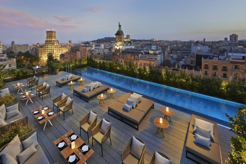 Top Barcelona Hotels with Rooftop Pool 19 Experience one of the finest Barcelona Hotels with Rooftop Pool, where you can unwind at sunset on a stylish terrace featuring a long pool, cushioned lounge seating, elegant dining tables, soft lighting, and stunning views of historic city buildings.