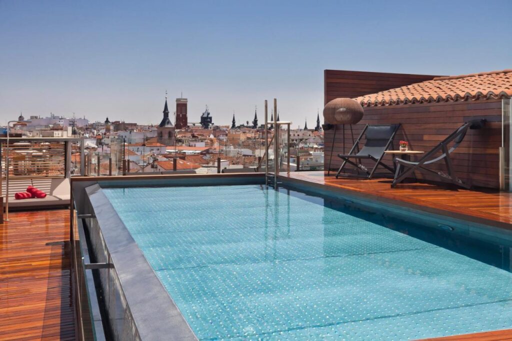 Where to Stay in Madrid: 14 Best Hotels 18 A rooftop pool with clear blue water overlooks a city skyline dotted with historic buildings—an ideal choice for where to stay in Madrid first time. Two lounge chairs, drinks on a table, and wooden decking create a relaxing urban retreat under the clear sky.