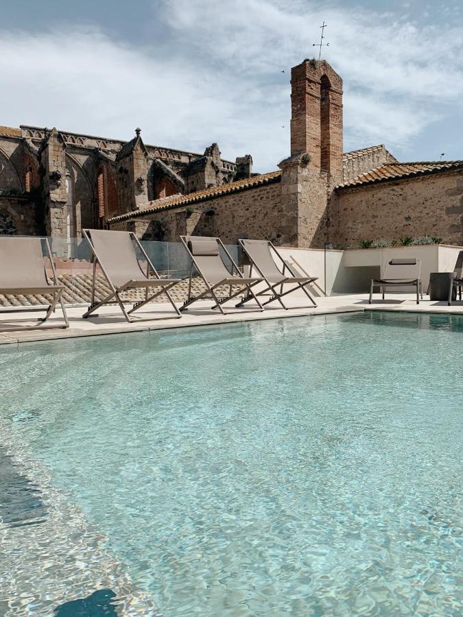 A clear swimming pool with lounge chairs beside it, set against an old stone building with a chimney and arched windows under a partly cloudy sky—perfect for those seeking boutique hotels in Costa Brava.