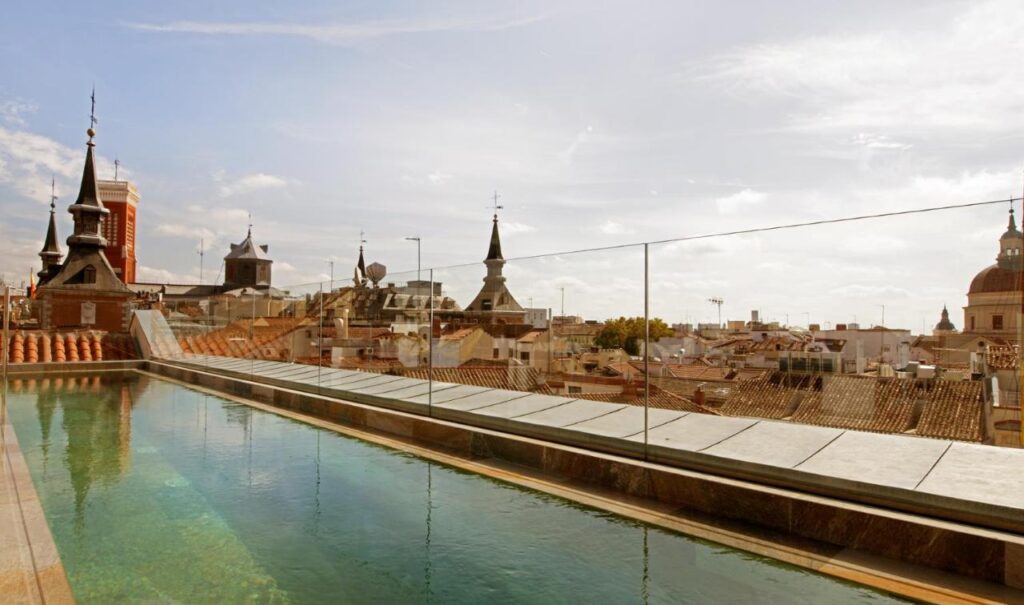 Where to Stay in Madrid: 14 Best Hotels 9 A rooftop swimming pool with clear water and glass fencing overlooks a cityscape of historic buildings, tiled rooftops, and church domes—an ideal spot when searching where to stay in Madrid first time.