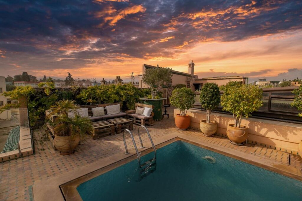 7 UNIQUE Riads in Fez You Won't Forget 27 A rooftop terrace inspired by riads in Fez, with a small swimming pool, potted plants, wooden benches with cushions, and a dramatic sunset sky with colorful clouds in the background.