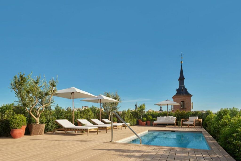 Where to Stay in Madrid: 14 Best Hotels 22 A rooftop pool area with sun loungers, white umbrellas, potted plants, and shrubs—an ideal spot when deciding where to stay in Madrid first time. A church tower rises in the background under a clear blue sky.