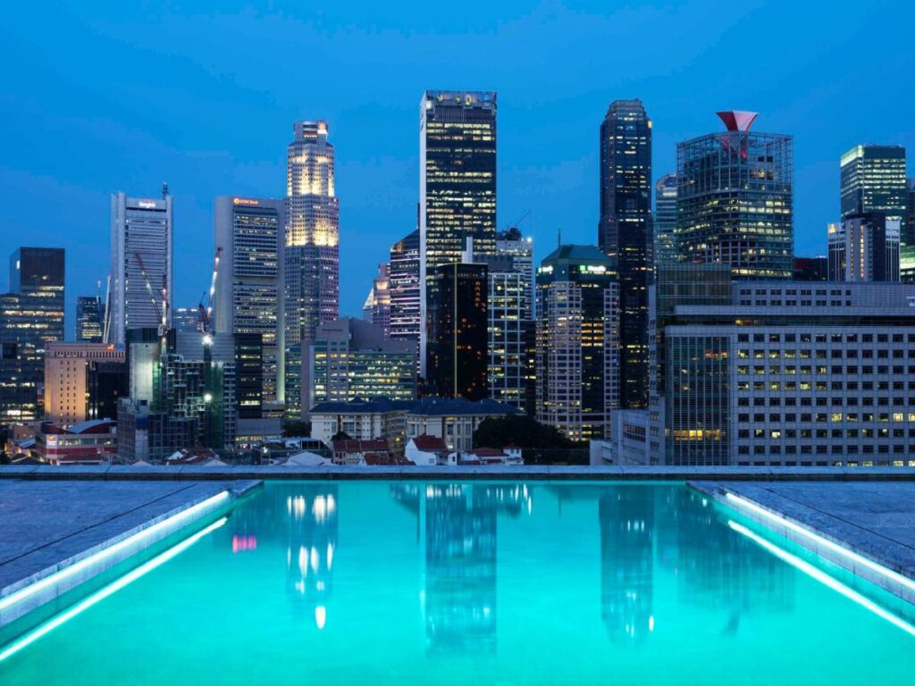 13 Most Unique Hotels in Singapore for an Incredible Holiday 48 A rooftop swimming pool glows with turquoise water in the foreground, reflecting the illuminated skyscrapers of a modern city skyline at dusk—an iconic scene at one of the unique hotels in Singapore.