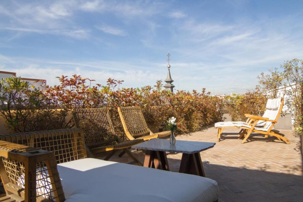 Where to Stay in Madrid: 14 Best Hotels 35 A sunny rooftop terrace with wicker chairs, a marble table, lounge chairs, and potted plants—an ideal spot for where to stay in Madrid first time—surrounded by greenery and blue sky. A church steeple rises in the background.