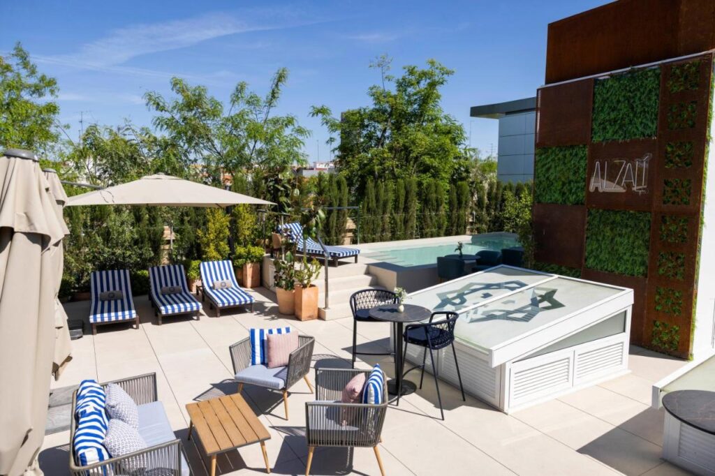 10 UNIQUE Hotels with Pool in Seville 12 A rooftop terrace in Seville featuring lounge chairs, striped cushions, umbrellas, a small pool, and greenery. Perfect for relaxing, this spot stands out among hotels with pool in Seville, with a sunny view and the word ALZÁI displayed on a plant wall.