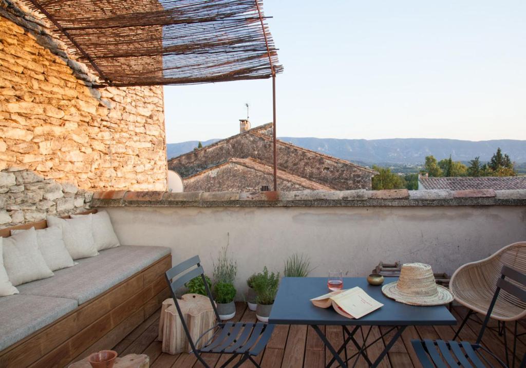 11 Unique Places to Stay in Provence 44 A cozy rooftop terrace with a wooden bench, cushions, a black table with drinks, a straw hat, chairs, potted plants, and rustic stone walls—one of the unique places to stay in Provence—overlooking rooftops and distant hills under a clear sky.
