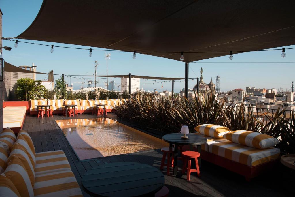Top Barcelona Hotels with Rooftop Pool 44 A stylish rooftop terrace at one of the top Barcelona Hotels with Rooftop Pool, featuring yellow and white striped lounge chairs, a shallow water feature, string lights overhead, and a city skyline under a canopy shade.