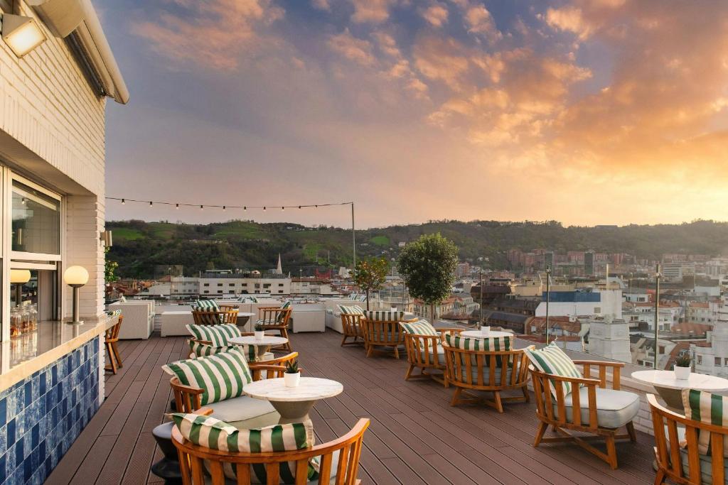 8 Unique Boutique Hotels in Bilbao Spain 15 Rooftop terrace at one of the boutique hotels in Bilbao, with round tables and cushioned wooden chairs, overlooks a sunset cityscape. Decorative string lights and greenery enhance the cozy, inviting atmosphere.