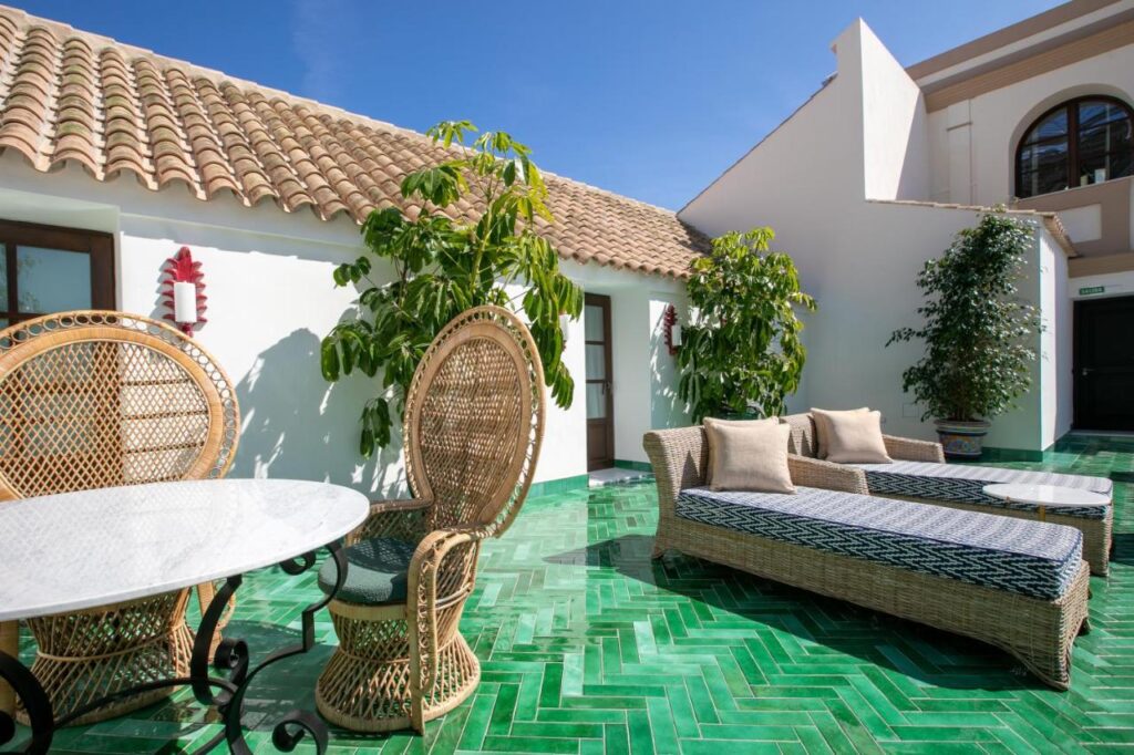 10 UNIQUE Hotels with Pool in Seville 19 Sunny patio with green herringbone tile floor, wicker chairs around a round white table, two loungers with cushions, and potted plants by white stucco walls—reminiscent of hotels with pool in Seville and their Mediterranean charm.