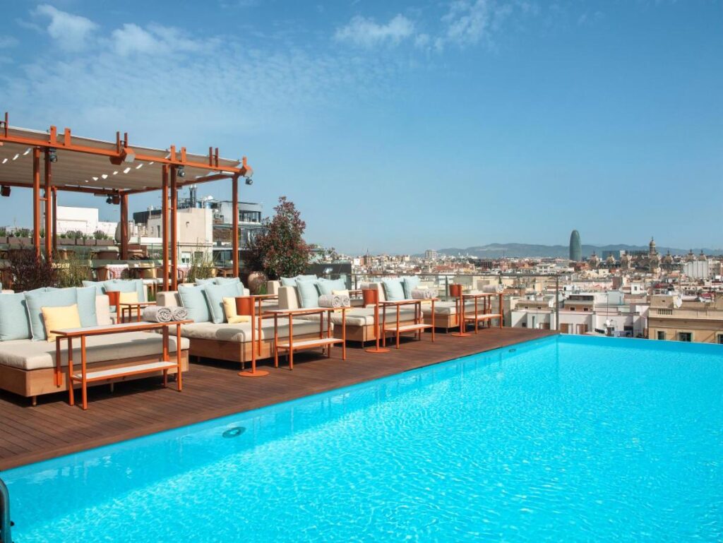 Top Barcelona Hotels with Rooftop Pool 22 A rooftop pool with clear blue water, surrounded by cushioned lounge chairs and sofas. The seating area offers classic views found in Barcelona Hotels with Rooftop Pool—cityscape vistas, bright skies, and hills in the distance.