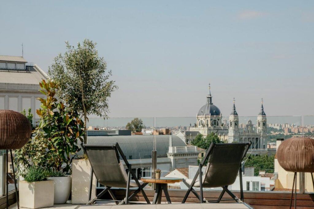 Where to Stay in Madrid: 14 Best Hotels 16 Two lounge chairs and a small table await on a rooftop terrace with potted plants, offering city rooftop views and a distant domed cathedral—an ideal spot for those wondering where to stay in Madrid first time.
