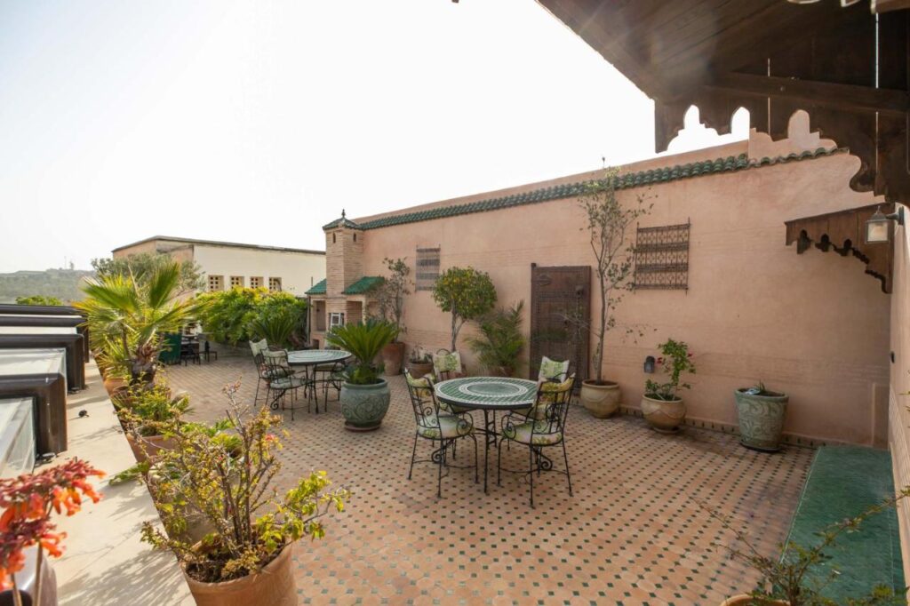 7 UNIQUE Riads in Fez You Won't Forget 30 A sunlit rooftop terrace, reminiscent of riads in Fez, features tiled flooring, potted plants, and several round tables with chairs set against a pale pink wall with green roof tiles and decorative accents.