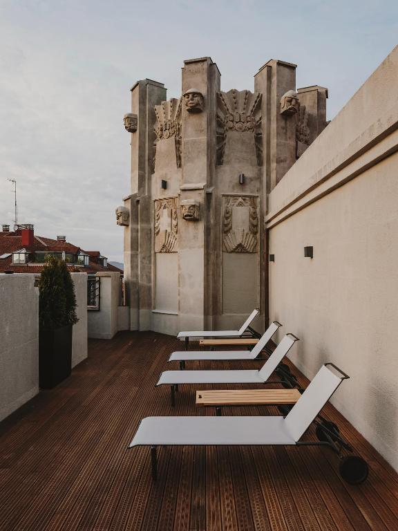Four white lounge chairs are arranged on a wooden rooftop deck, set against a tall, ornate stone structure with sculpted faces. The scene evokes the elegance of boutique hotels in Asturias, with cloudy skies and city buildings in the background.